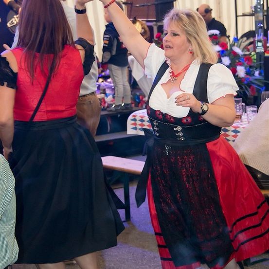 Eine Frau in einem traditionellen deutschen Kleid tanzt mit einer anderen Frau bei einer lebhaften Veranstaltung, mit einer Band, die im Hintergrund spielt.