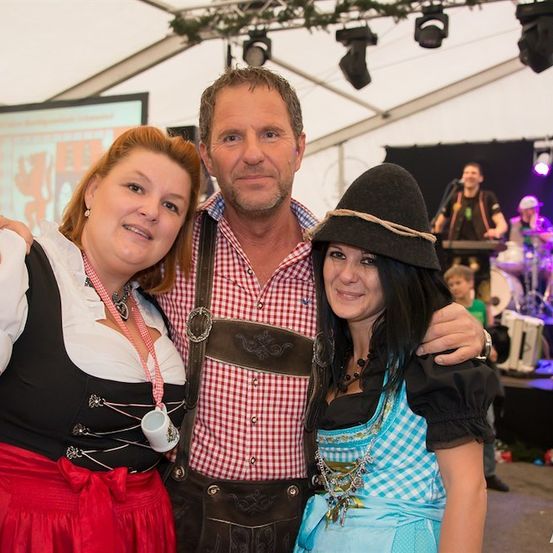 Drei Menschen lächeln und posieren für ein Foto bei einer festlichen Veranstaltung, wobei ein Mann ein kariertes Hemd und Hosenträger trägt und zwei Frauen in traditioneller Kleidung. Im Hintergrund ist eine Bühne mit Musikinstrumenten und einem Künstler zu sehen.