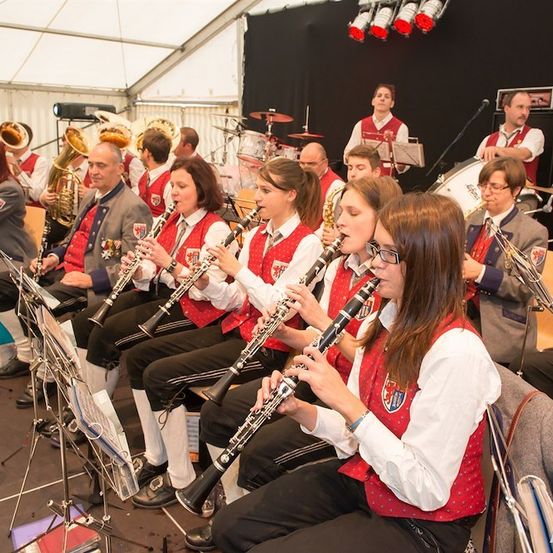 Eine Gruppe von Musikern in roten Westen und weißen Hemden spielt ihre Instrumente, einschließlich Klarinetten, bei einer Aufführung. Sie sitzen auf Stühlen und haben Notenständer vor sich. Dahinter spielen andere Musiker verschiedene Instrumente. Die Bühne ist gut beleuchtet mit Scheinwerfern.