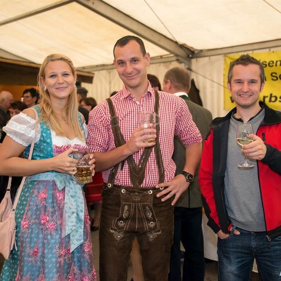 Drei Menschen stehen in einem Zelt, lächeln und halten Getränke in der Hand. Die Frau trägt ein traditionelles Kleid, und die Männer sind in Freizeitkleidung. Sie sind von anderen Menschen umgeben, und im Hintergrund befindet sich ein gelbes Banner.