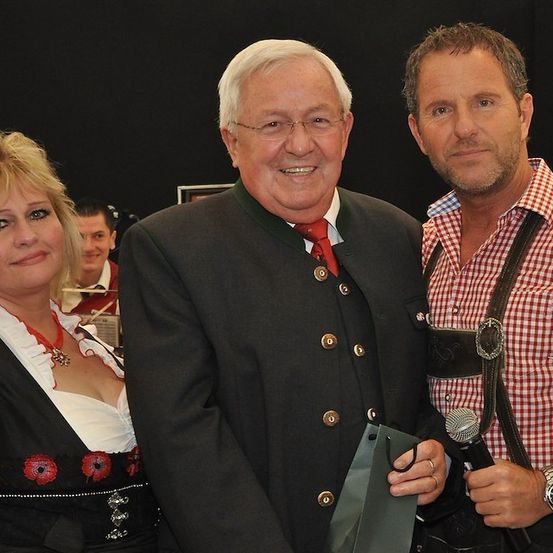 Drei Menschen, zwei Männer und eine Frau, lächeln und posieren für ein Foto. Der Mann links trägt einen Anzug und eine Krawatte, die Frau ein traditionelles Kleid und der Mann rechts ein kariertes Hemd und eine Hose mit Aufhängung.