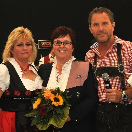 Drei Menschen stehen eng beieinander und lächeln. Eine Frau auf der linken Seite hält einen Blumenstrauß. Die Frau in der Mitte trägt eine Brille. Der Mann auf der rechten Seite hält ein Mikrofon.