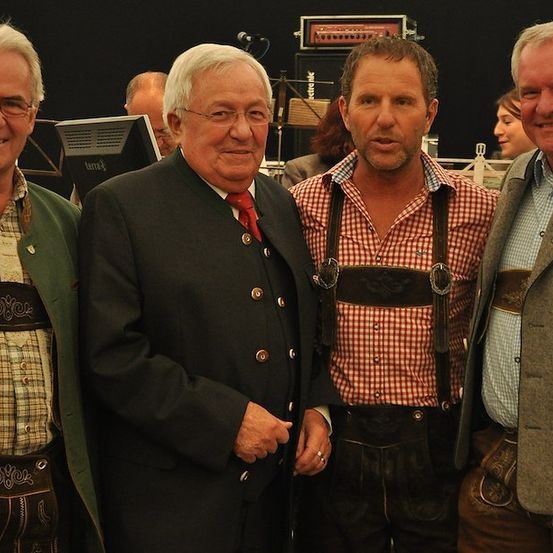Vier Männer in traditioneller deutscher Kleidung stehen zusammen und lächeln für ein Foto. Der Mann in der Mitte trägt ein rot-weiß kariertes Hemd und eine Lederhose. Im Hintergrund ist eine verschwommene Kulisse mit einem schwarzen Hintergrund und einem Computermonitor zu sehen.
