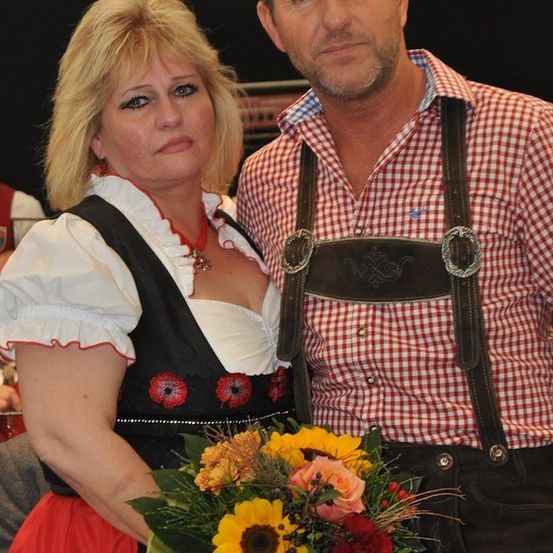 Ein Mann und eine Frau in traditioneller deutscher Kleidung, die eng beieinanderstehen und einen Blumenstrauß halten. Die Frau trägt ein rot-schwarzes Kleid mit roten Blumen, während der Mann ein rot-weiß kariertes Hemd mit Hosenträger trägt.
