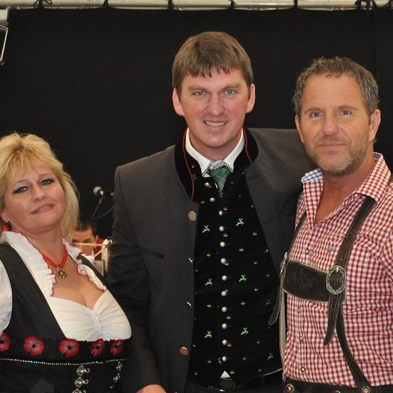Drei Menschen in traditioneller deutscher Kleidung stehen zusammen und lächeln für ein Foto. Der Mann in der Mitte trägt eine grüne Krawatte und eine schwarze Weste mit dekorativen Knöpfen. Die Frau links trägt ein weißes und schwarzes Kleid mit roten Blumen. Der Mann rechts trägt ein rot-weiß kariertes Hemd und eine Hose mit Trägern.