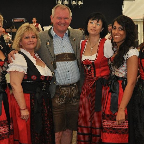 Eine Gruppe von Menschen in traditioneller bayerischer Kleidung, lächelnd und posierend für ein Foto. Der Mann in der Mitte trägt ein blaues Hemd und braune Shorts.