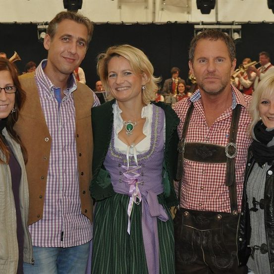 Fünf Personen posieren für ein Foto, mit einer Frau in traditioneller bayerischer Kleidung in der Mitte. Dahinter spielt eine Band, und eine Menge schaut zu.