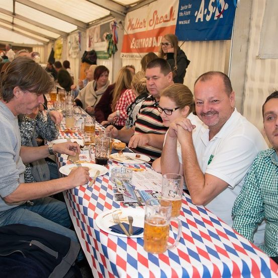 Eine Gruppe von Menschen sitzt an einem langen Tisch und genießt Essen und Getränke. Der Tisch ist mit einer rot-weiß-blauen Tischdecke bedeckt. Einige der Leute lächeln und schauen in die Kamera. Auf dem Tisch stehen mehrere Gläser Bier. Im Hintergrund befindet sich ein Banner mit dem Text 'Kelter Speak'.