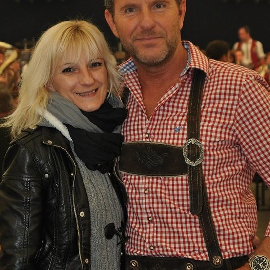 Ein Mann und eine Frau lächeln und posieren für ein Foto auf einem Festival. Der Mann trägt traditionelle bayerische Kleidung, während die Frau in einer schwarzen Lederjacke gekleidet ist.
