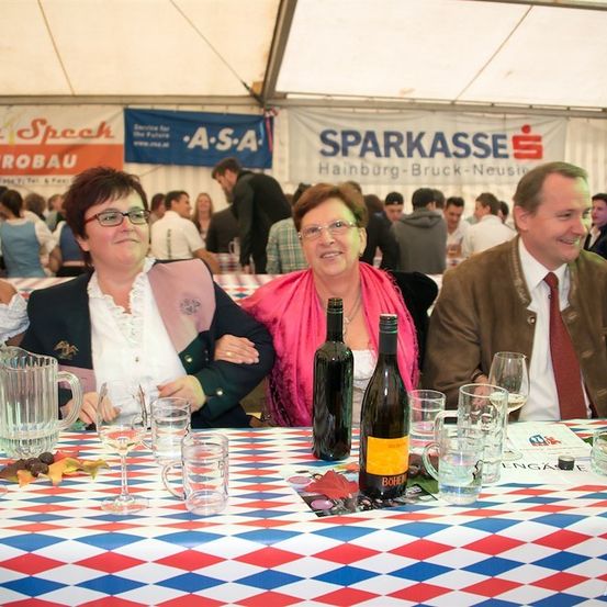 Drei Personen sitzen an einem Tisch mit Weingläsern und Flaschen und lächeln für die Kamera. Ein rot-weiß-blauer Tischläufer ist sichtbar, und ein Banner hinter ihnen trägt die Aufschrift 'SPARKASSE'.
