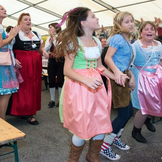 Eine Gruppe von Frauen in traditioneller deutscher Kleidung tanzt und singt im Freien, mit einer Holzbank und einem Zelt im Hintergrund.