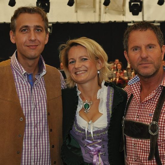 Drei Menschen stehen bei einem Event zusammen. Der Mann links trägt eine braune Weste, die Frau in der Mitte ein traditionelles Kleid und der Mann rechts ein kariertes Hemd mit Hosenträgern. Sie alle lächeln.