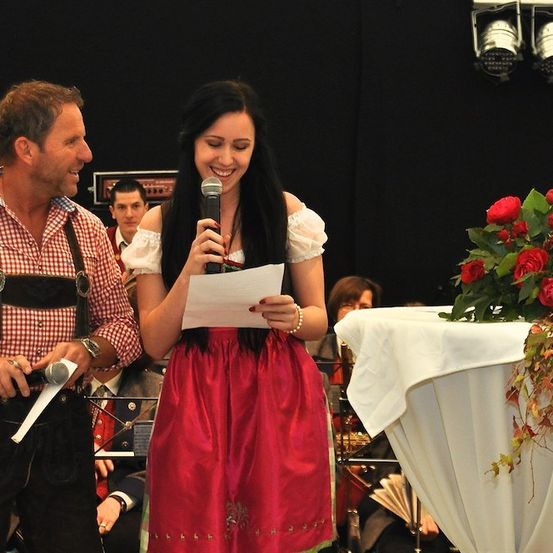 Ein Mann und eine Frau stehen auf der Bühne, die Frau hält ein Mikrofon und ein Stück Papier, lächelt und schaut den Mann an, der auch lächelt. Dahinter steht ein Tisch mit einer Blumenarrangement und einem schwarzen Hintergrund.