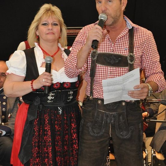 Ein Mann und eine Frau in traditioneller deutscher Kleidung stehen und halten Mikrofone, möglicherweise bei einer Bühnenaufführung.