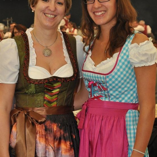 Zwei Frauen in traditioneller bayerischer Kleidung, eine in einem grünen Kleid und die andere in einem blau-weiß karierten Kleid, lächeln für die Kamera.
