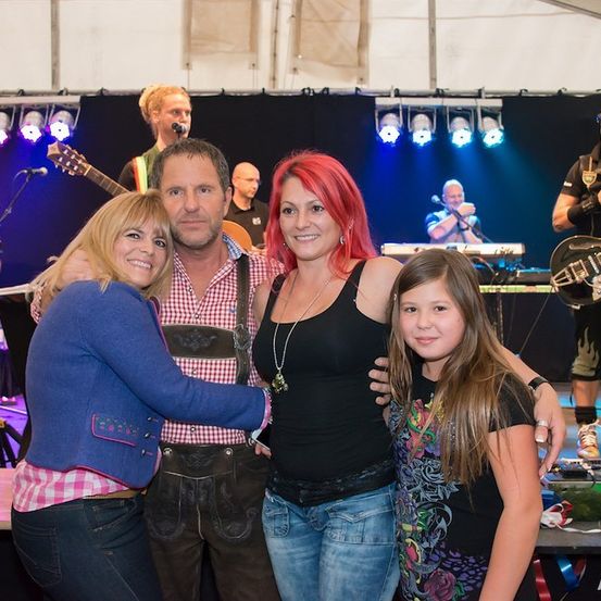 Eine Gruppe von Menschen posiert für ein Foto auf einer Bühne, mit einer Band im Hintergrund. Die Band spielt Instrumente und singt.