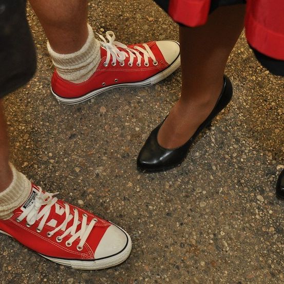 Drei Personen stehen auf einer Kiesoberfläche. Die Person auf der linken Seite trägt rote Converse-Sneaker. Die Person in der Mitte trägt schwarze Schuhe. Die Person auf der rechten Seite trägt einen roten Mantel.