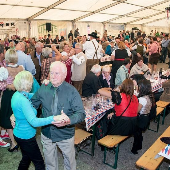 Eine große Gruppe von Menschen versammelt sich in einem Zelt für eine festliche Veranstaltung, einige tanzen, während andere an Tischen mit Essen und Getränken sitzen. Das Zelt ist mit Bannern und Flaggen geschmückt.
