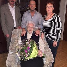 Eine ältere Frau sitzt in einem Stuhl und hält einen Blumenstrauß, mit zwei Männern und einer Frau hinter ihr.