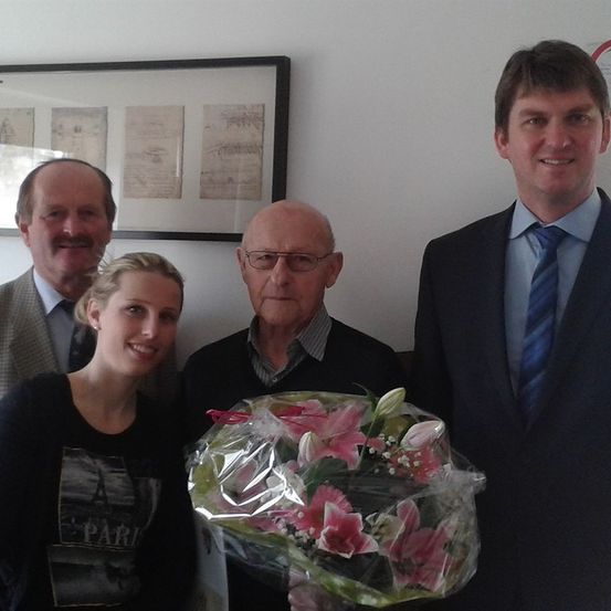 Vier Personen stehen in einem Raum mit einer weißen Wand und einem gerahmten Bild. Ein Mann hält einen Blumenstrauß.