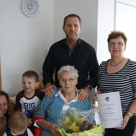 Ein Familienfoto mit einer älteren Frau, die einen Blumenstrauß und ein Zertifikat hält, steht neben einem Mann und einer Frau, und ein junger Junge und ein Mädchen lächeln im Hintergrund.