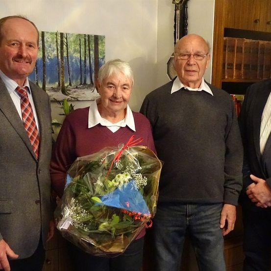 Vier Personen stehen in einem Raum, zwei Männer und zwei Frauen. Eine Frau hält einen Blumenstrauß. Die Männer tragen Anzüge und Krawatten.