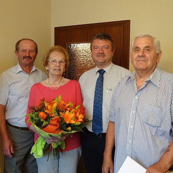 Vier Personen, drei Männer und eine Frau, stehen zusammen vor einer Holztür. Die Frau hält einen Blumenstrauß.