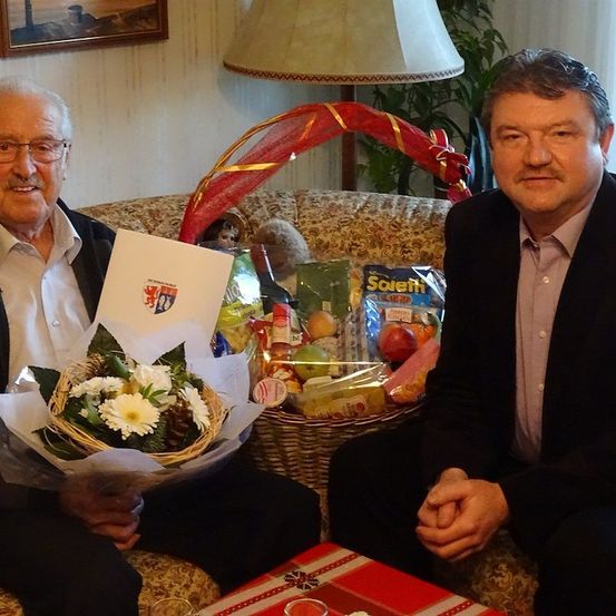 Zwei Männer sitzen auf einer Couch, einer hält einen Blumenstrauß und eine Karte, der andere mit ernster Miene. Dahinter steht ein Korb mit verschiedenen Gegenständen und eine Lampe.