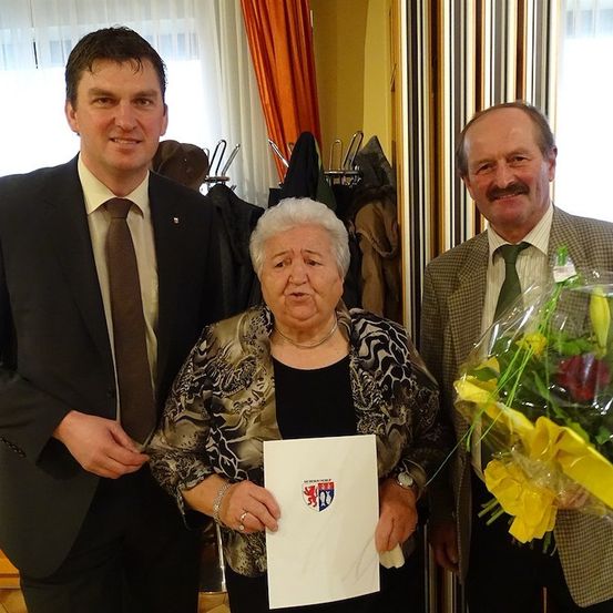 Drei Menschen stehen lächelnd zusammen. Eine Frau hält ein Papier mit einem Wappen, und ein Mann auf der rechten Seite hält einen Blumenstrauß.