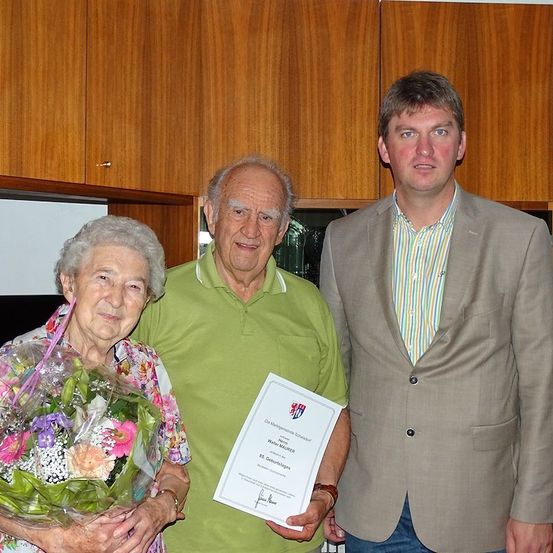 Eine ältere Frau mit einem Blumenstrauß steht zwischen zwei Männern, von denen einer ein Zertifikat in der Hand hält. Der Mann auf der rechten Seite trägt einen Anzug, während der Mann auf der linken Seite ein grünes Poloshirt trägt. Sie stehen vor Holzschränken.