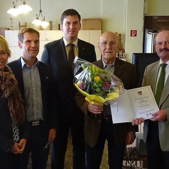 Fünf Personen stehen lächelnd in einem Raum. Der Mann in der Mitte hält einen Blumenstrauß und ein Zertifikat. Hinter ihnen befinden sich Schränke und eine Lampe.