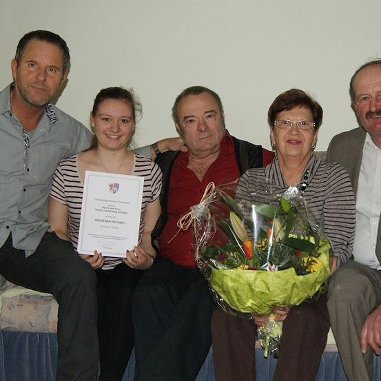 Eine Familie von fünf posiert für ein Foto mit einer Frau, die einen Blumenstrauß und ein Zertifikat hält. Der Mann links trägt eine Halskette und eine Uhr. Die Frau in der Mitte hält ein Zertifikat und lächelt. Der Mann rechts trägt eine Brille.
