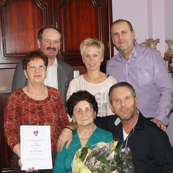 Eine Familie von fünf Erwachsenen, zwei Frauen und drei Männern, posiert für ein Foto. Die Frau auf der linken Seite hält eine Urkunde, und die Frau in der Mitte hält einen Blumenstrauß. Der Mann auf der rechten Seite lächelt und der Mann auf der linken Seite schaut in die Kamera. Hinter ihnen steht ein Holzschrank mit einer Lampe.