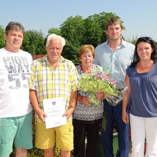 Eine Gruppe von Menschen steht draußen und lächelt. Ein Mann hält ein Zertifikat, während eine Frau Blumen hält. Dahinter hält eine Frau ein Zertifikat und ein Mann trägt eine Sonnenbrille auf dem Kopf.