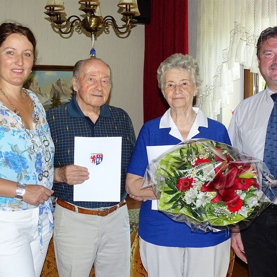 Vier Personen stehen lächelnd für ein Foto zusammen im Innenraum. Ein älterer Mann hält ein Zertifikat, während eine ältere Frau einen Blumenstrauß hält. Ein Mann und eine Frau stehen auf beiden Seiten von ihnen.