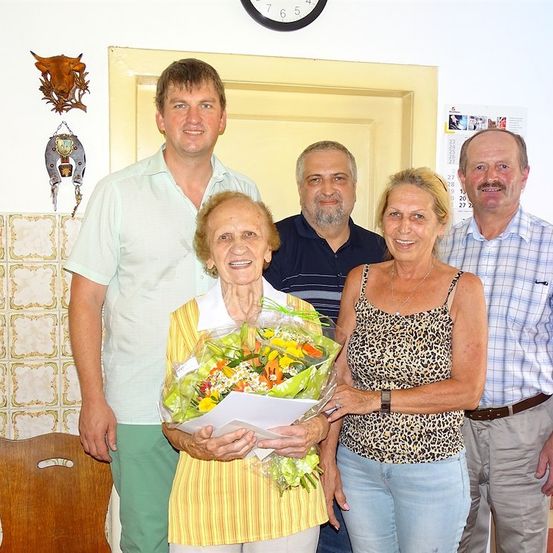 Eine Gruppe von Menschen steht in einem Raum, eine Frau hält einen Blumenstrauß. Hinter ihnen befindet sich ein Kalender an der Wand und eine Uhr.