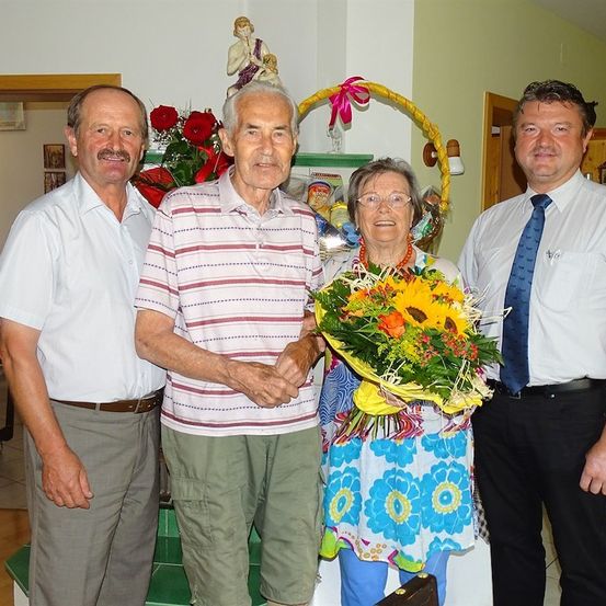 Vier Menschen stehen zusammen in einem Raum und lächeln für ein Foto. Eine Frau hält einen Blumenstrauß. Hinter ihnen ist eine Wand mit einer Statue und einem Korb mit Blumen.