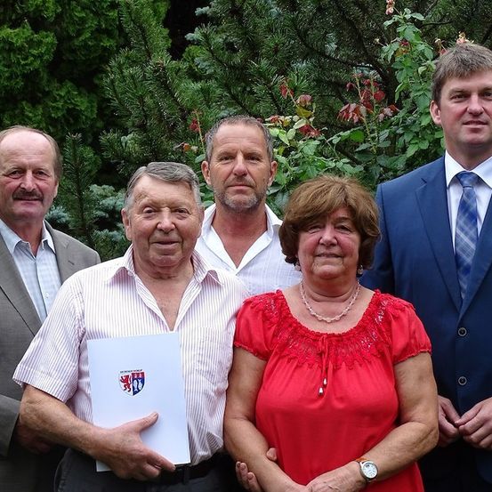 Fünf Personen, drei Männer und zwei Frauen, stehen gemeinsam in einem Garten und lächeln für ein Foto. Der Mann in der Mitte hält ein Papier mit einem Logo.