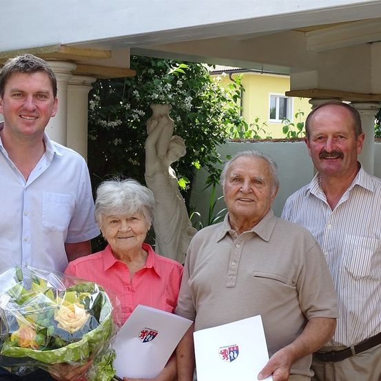 Drei Menschen stehen lächelnd unter einem Vordach. Die Frau hält einen Blumenstrauß und ein Zertifikat. Der Mann rechts trägt einen Gürtel.
