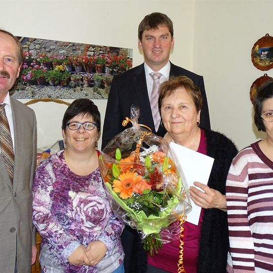Fünf Menschen stehen lächelnd in einem Raum. Eine Frau hält einen Blumenstrauß und ein Papier. Hinter ihnen ist eine Wand mit einem Bild von Blumen.