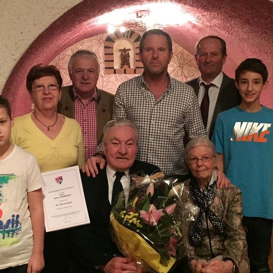 Eine Gruppe von Menschen ist zu einer Feier versammelt, wobei ein älterer Mann einen Blumenstrauß und ein Zertifikat in der Hand hält. Eine ältere Frau sitzt neben ihm. Hinter ihnen steht ein Mann im Anzug mit einem jüngeren Jungen.