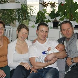 Eine vierköpfige Familie sitzt auf einer Couch und lächelt für ein Foto. Der Mann in der Mitte hält ein Baby. Hinter ihnen stehen Topfpflanzen und ein weißes Kinderbett.