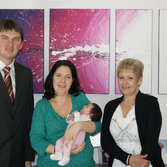 Ein Mann und zwei Frauen stehen vor einer Wand mit abstrakten Gemälden. Die Frau in der Mitte hält ein Baby.