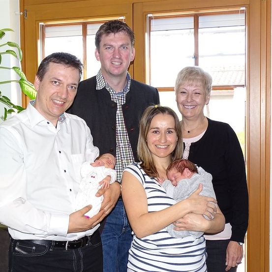 Eine Familie mit vier Erwachsenen und zwei Neugeborenen posiert für ein Foto vor einem Fenster. Der Mann links hält ein Baby, während die Frau in der Mitte ein weiteres hält. Die anderen beiden Erwachsenen stehen hinter ihnen.