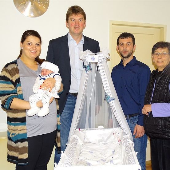 Eine vierköpfige Familie steht um ein Kinderbett mit einem Baby darin und lächelt für ein Foto. Die Mutter hält das Baby, während der Vater und zwei andere in der Nähe stehen.