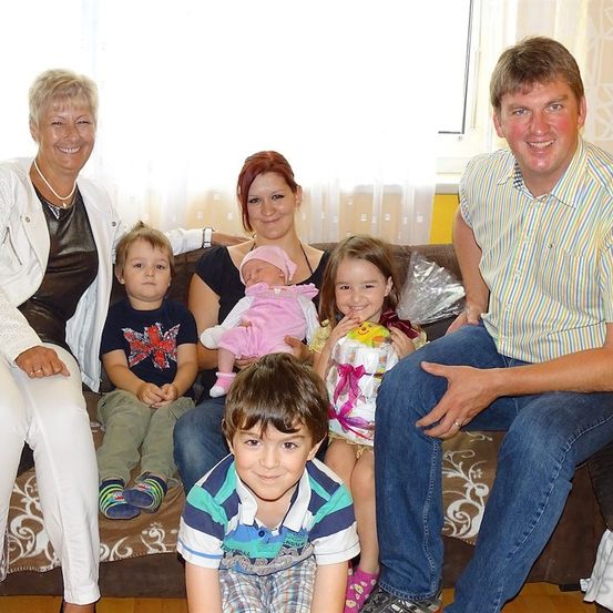 Eine Familie mit fünf Personen sitzt auf einem Sofa und lächelt für ein Foto. Die Frau in der Mitte hält ein Baby. Der Mann rechts trägt ein gestreiftes Hemd.