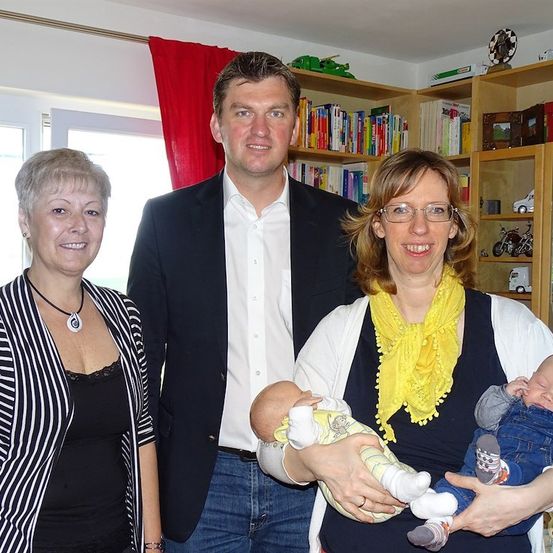 Ein Familienfoto mit zwei Frauen und einem Mann, alle lächelnd, vor einem Bücherregal mit Büchern und Spielzeug. Die Frau rechts hält zwei Babys.