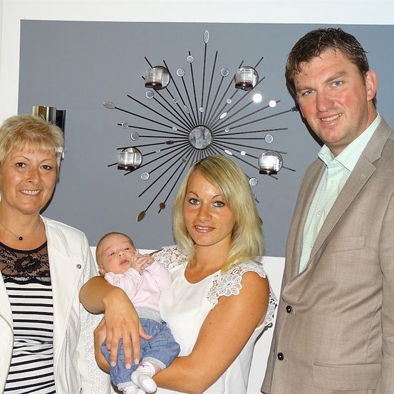 Ein Familienfoto mit einer Frau, die ein Baby hält und lächelt, neben einem Mann und einer weiteren Frau. Sie stehen vor einer grauen Wand mit einem dekorativen Metall-Sonnenblume.