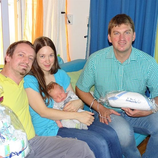 Eine Familie mit drei Erwachsenen und einem Neugeborenen sitzt lächelnd auf einer Couch. Der Mann rechts hält ein verpacktes Geschenk.