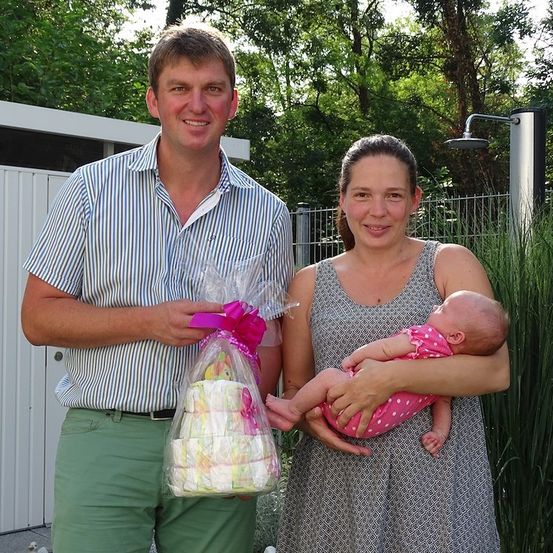 Ein Mann und eine Frau stehen draußen mit einem Baby. Der Mann hält eine verpackte Torte und die Frau ein Baby in einem rosa gepunkteten Outfit. Sie lächeln.